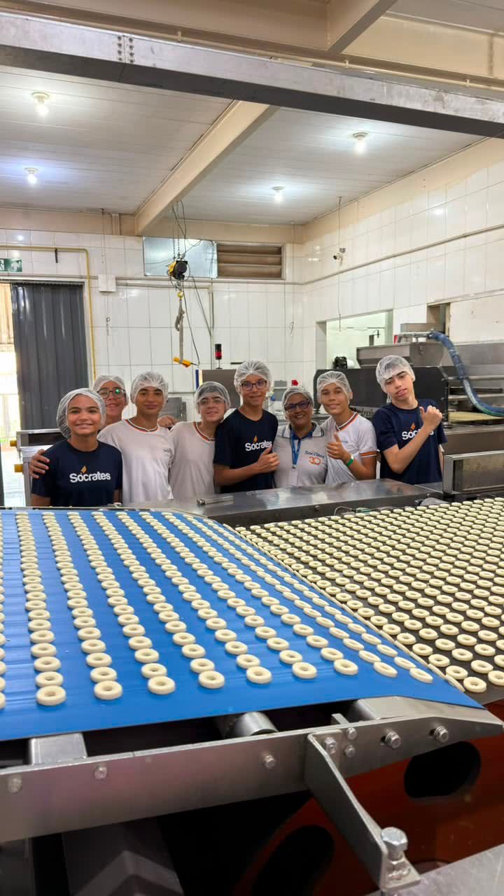 Alunos do Colégio Sócrates de touca ao lado da linha de produção de biscoitos