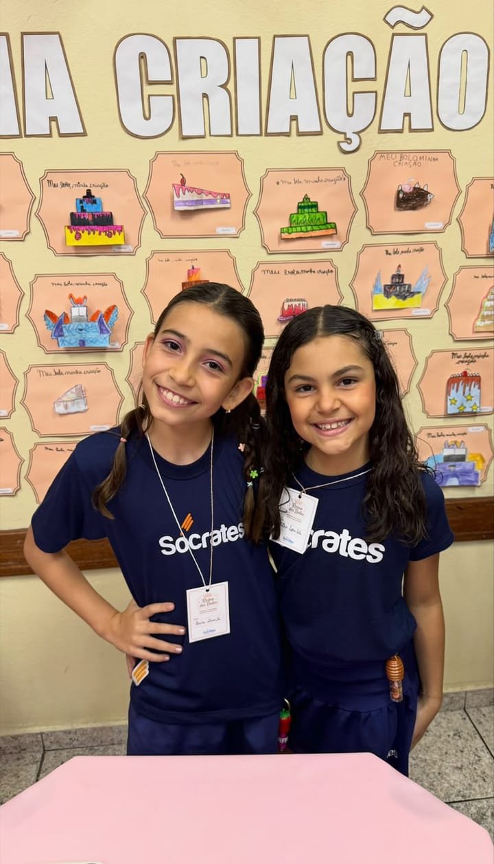 Duas alunas sorrindo em frente ao painel de bolos desenhados pela turma