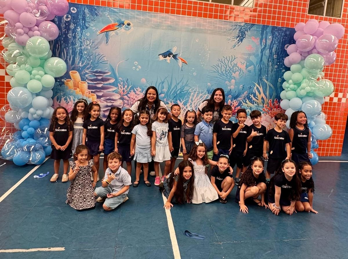 Crianças da Educação Infantil do Colégio Sócrates reunidas na quadra em frente ao cenário de fundo do mar com arcos de balões