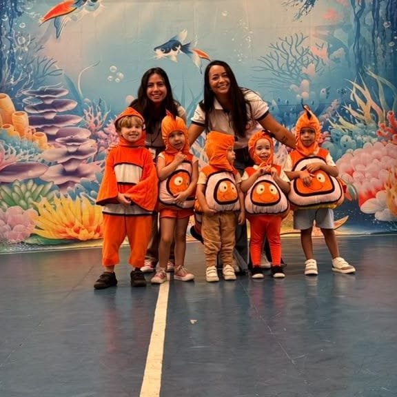 Crianças fantasiadas de peixe-palhaço com duas educadoras em frente ao cenário de fundo do mar